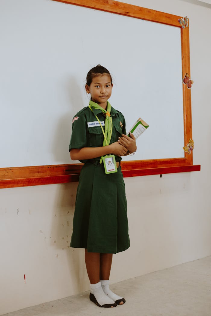 A young student in a green uniform stands confidently by a classroom whiteboard.