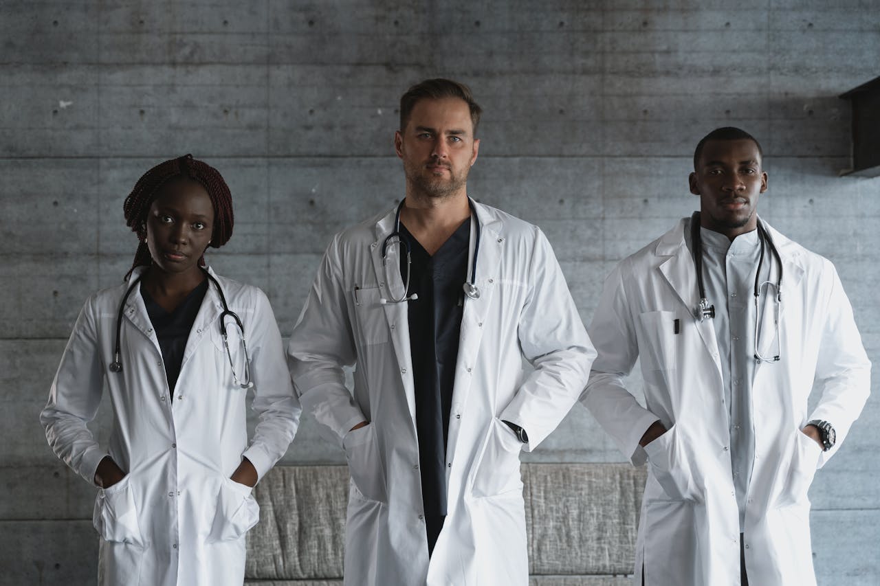 Three doctors in medical gowns with stethoscopes standing confidently indoors.